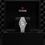 A silver TUDOR Royal wristwatch with a metal bracelet stands upright in a black presentation box, showcasing the TUDOR logo and name inside the lid—an elegant example of sport-chic timepieces.