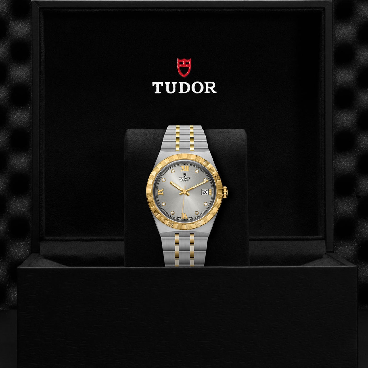 A TUDOR Royal wristwatch with a silver and gold integrated bracelet is presented in an open black box, featuring the TUDOR logo and name above the watch.