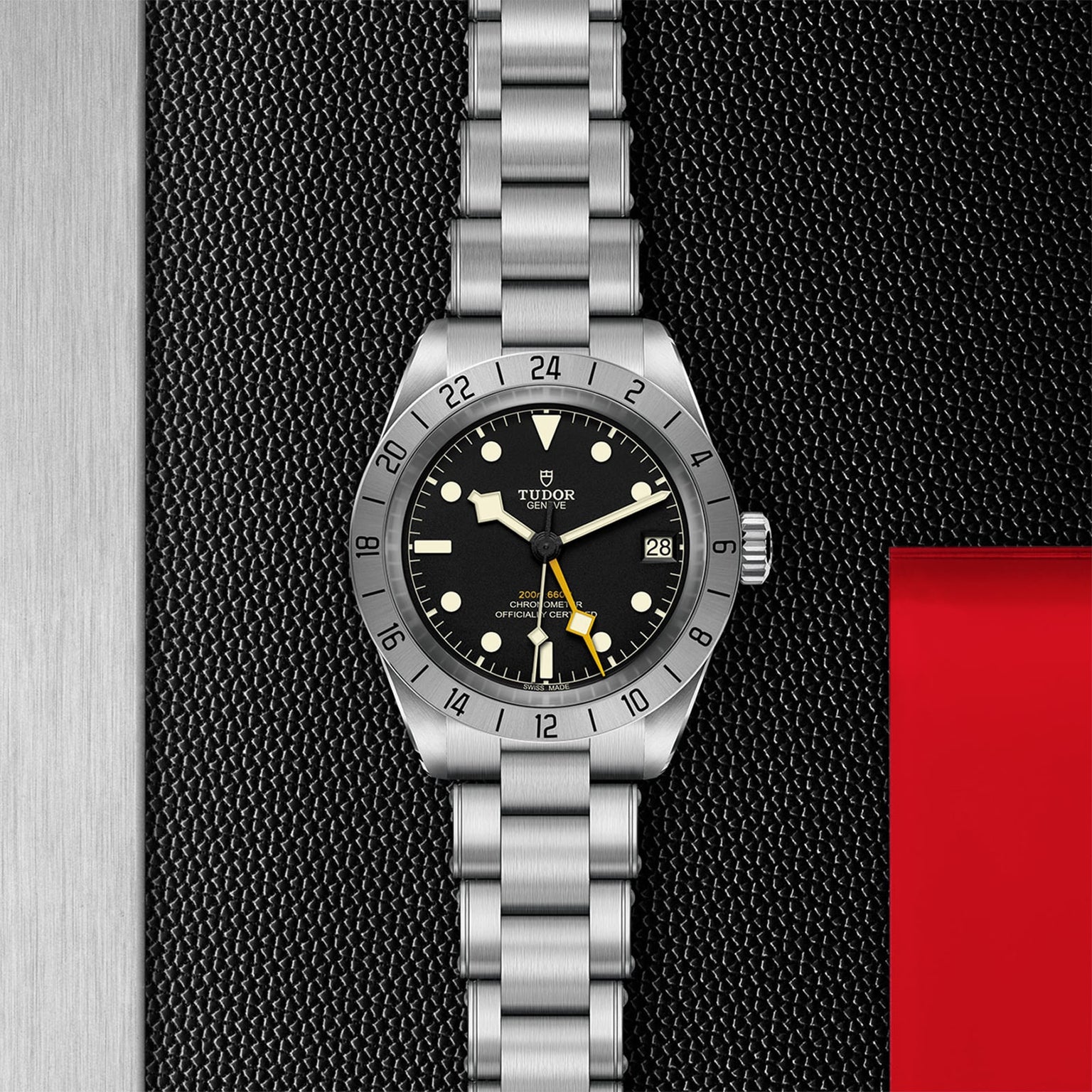 A TUDOR Black Bay Pro stainless steel wristwatch with a black dial, 24-hour bezel, and date display rests on a textured black surface beside a red accent, highlighting the brand’s heritage in modern watchmaking.