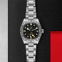 A TUDOR Black Bay Pro stainless steel wristwatch with a black dial, 24-hour bezel, and date display rests on a textured black surface beside a red accent, highlighting the brand’s heritage in modern watchmaking.