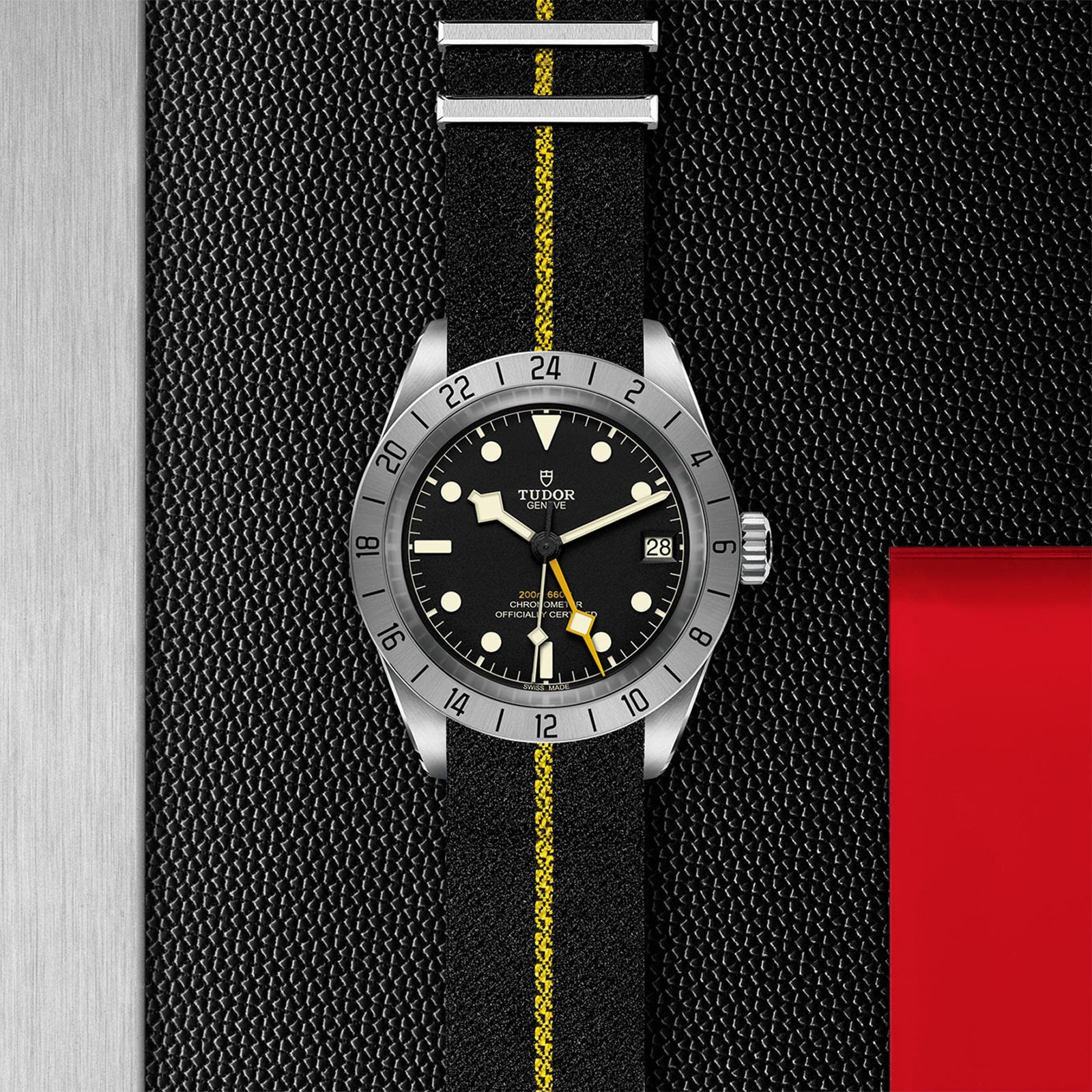 The TUDOR Black Bay Pro features a black and yellow striped fabric strap, black dial, date display, and silver bezel—displayed on a textured black-metal surface with red accent—ideal for Black Bay fans.