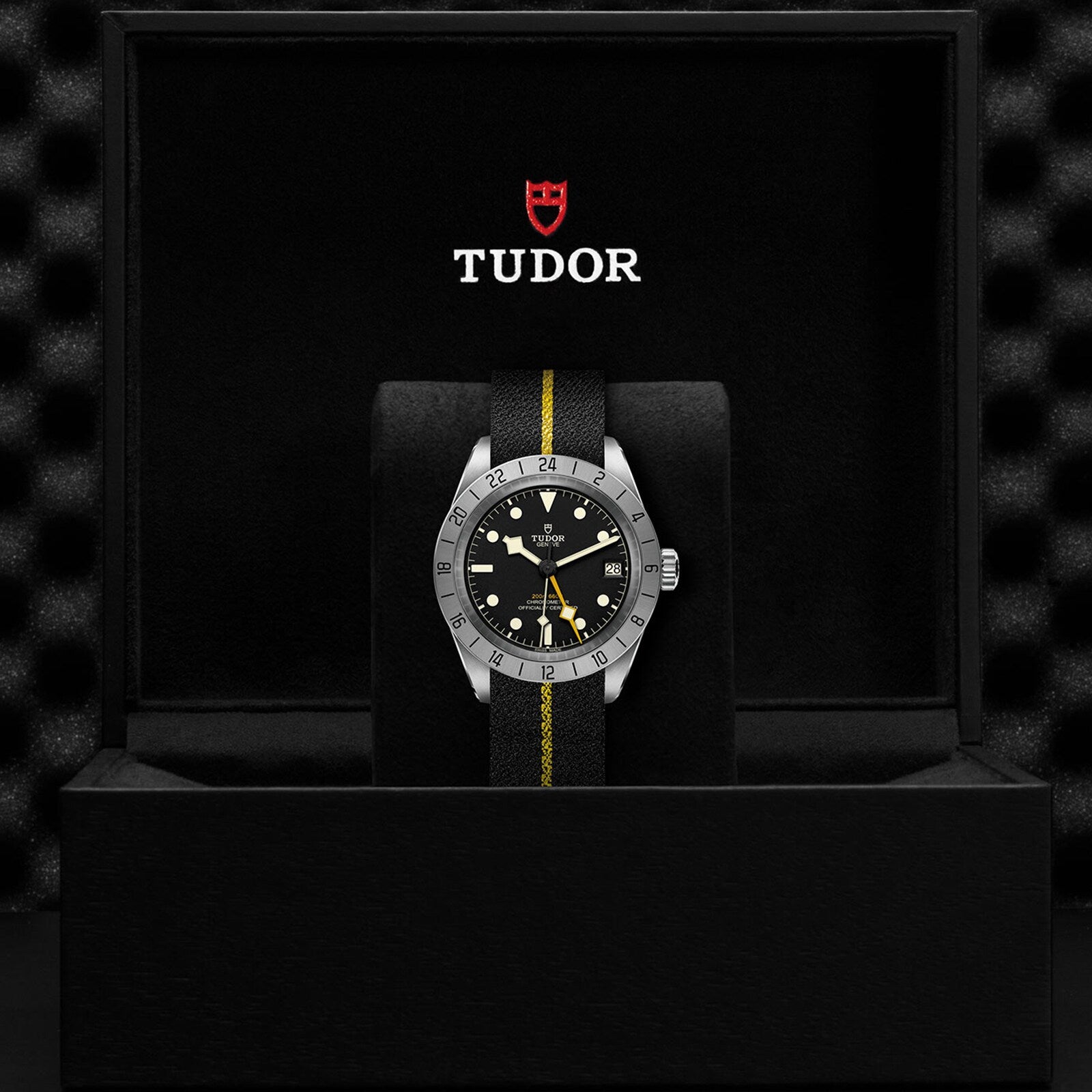 A neo-vintage TUDOR Black Bay Pro wristwatch with a black and yellow strap is displayed in an open black box featuring the TUDOR logo inside, reflecting the enduring legacy of TUDOR divers’ watches.