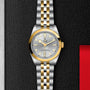 A TUDOR Black Bay One 31 S&G wristwatch with a silver dial, gold bezel, and steel-and-gold bracelet is displayed on a textured black background beside a red accent.