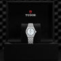 A TUDOR Royal wristwatch with a silver metal band and white face is displayed in an open black TUDOR-branded box.