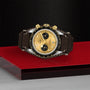 A TUDOR Black Bay Chrono S&G, featuring a gold and black chronograph dial and brown leather strap, is displayed on a red and gray platform against a textured black background.