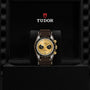 The TUDOR Black Bay Chrono S&G wristwatch, featuring a gold and black dial and brown strap, is elegantly displayed upright in a black Tudor-branded box—a refined addition to your automatic sport-chic collection.