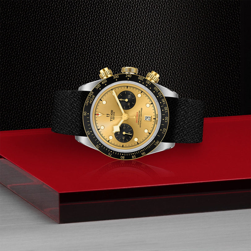 The TUDOR Black Bay Chrono S&G, a gold and black wristwatch with two subdials, black bezel, and black strap, is displayed on a red and gray surface against a black textured background.