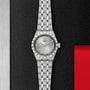 A TUDOR Royal wristwatch with a light gray dial and stainless steel and gold bracelet rests on a textured black surface next to a red and metal section.