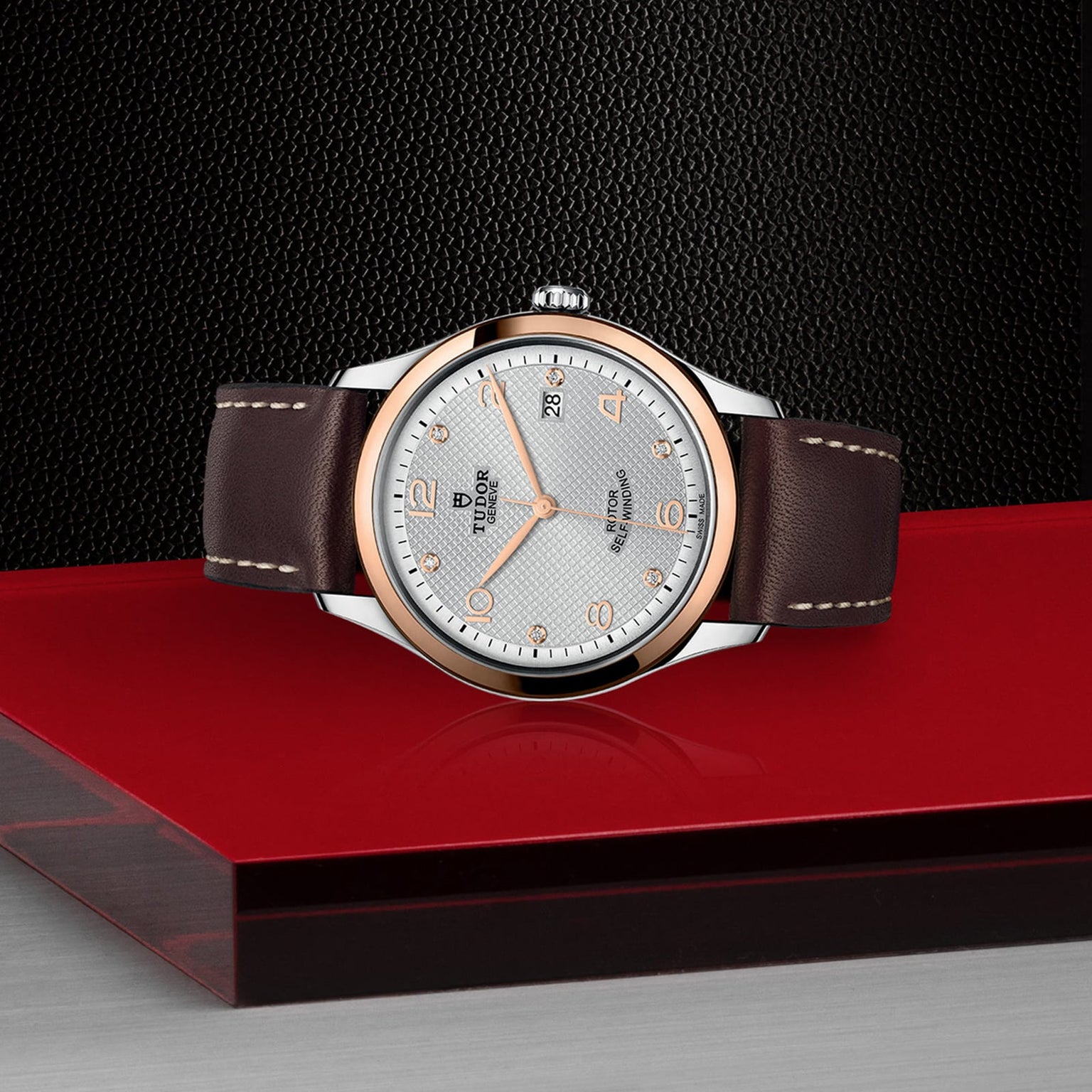 A TUDOR 1926 wristwatch featuring a brown leather strap, silver dial, gold accents, and date display is displayed on a red and gray surface with a black textured background.