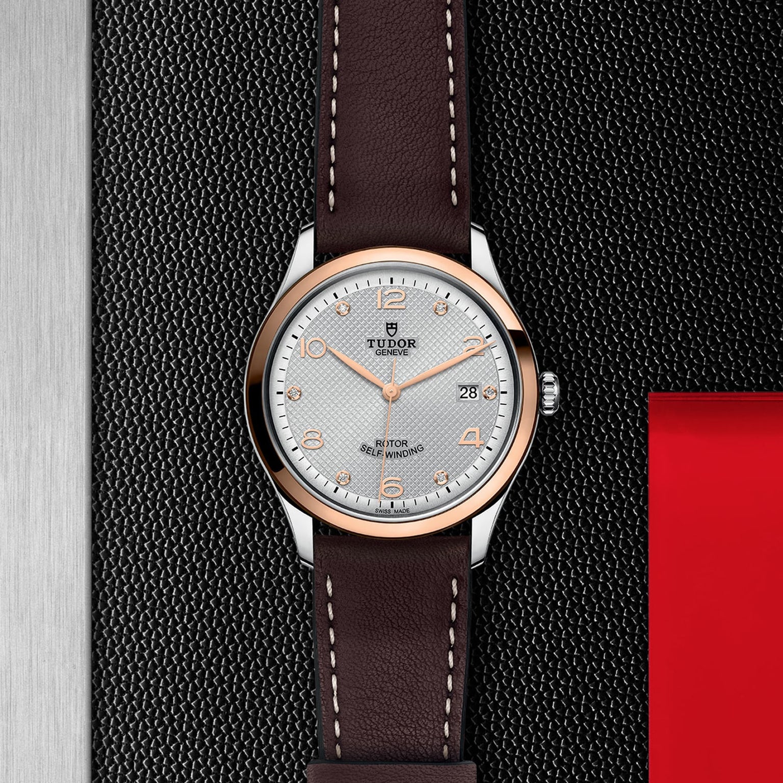 The TUDOR 1926 wristwatch, featuring a brown leather strap and silver dial, is displayed on a textured black surface.