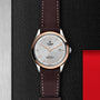 The TUDOR 1926 wristwatch, featuring a brown leather strap and silver dial, is displayed on a textured black surface.