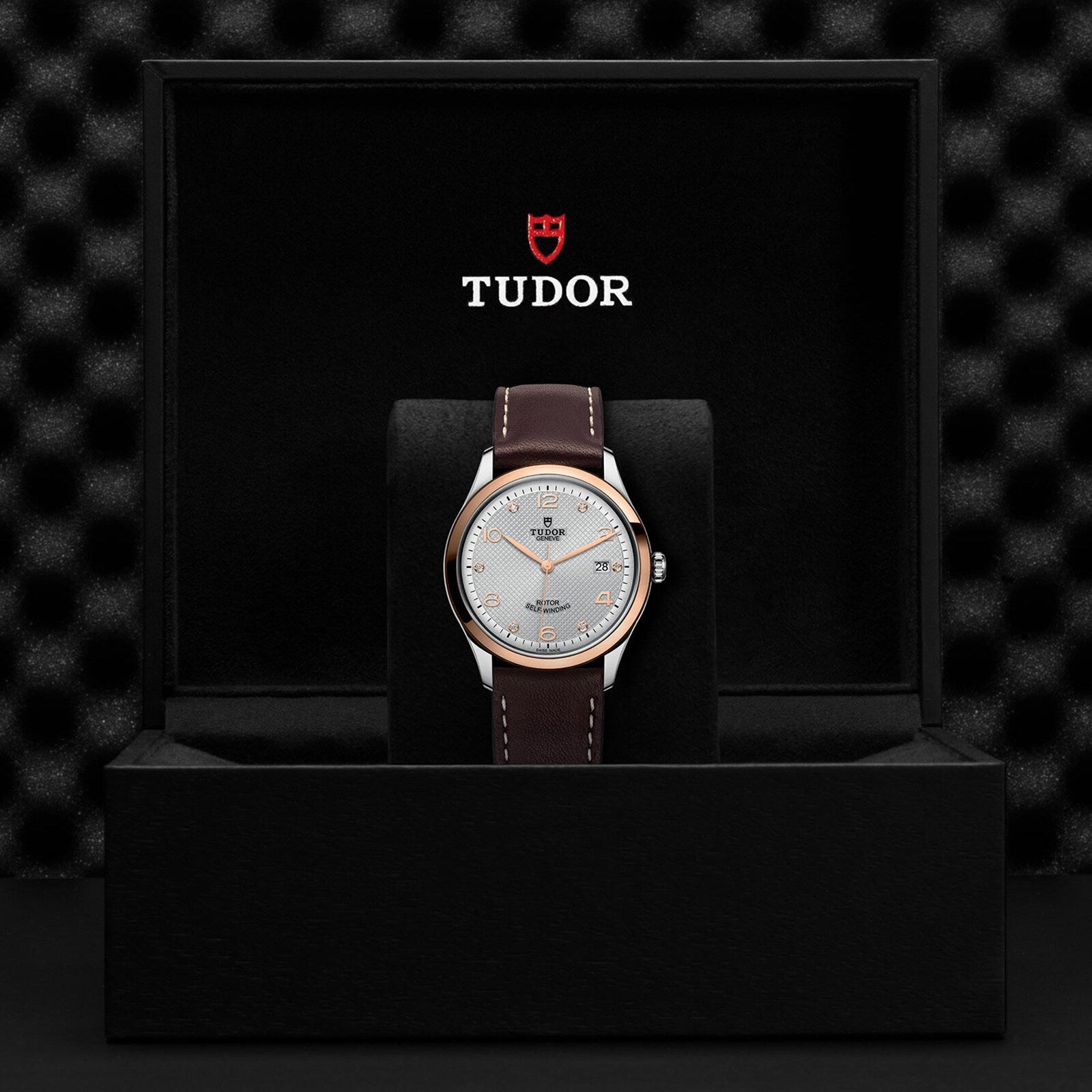 A TUDOR 1926 wristwatch with a brown leather strap and light dial is shown upright in a black presentation box, displaying the Tudor logo on the inside lid.