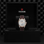 A TUDOR 1926 wristwatch with a brown leather strap and light dial is shown upright in a black presentation box, displaying the Tudor logo on the inside lid.