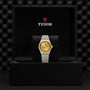 A TUDOR Royal wristwatch featuring a gold face and a stainless steel and gold bracelet is displayed in an open black Tudor box against a textured background.