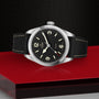 The TUDOR Ranger wristwatch, featuring Super Luminova on its black dial, silver bezel, and black textured strap, is displayed against a bold black and red geometric backdrop.