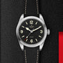 A TUDOR Ranger wristwatch featuring a black dial, beige Super Luminova numerals, and a black textured strap is showcased on a dark textured background.