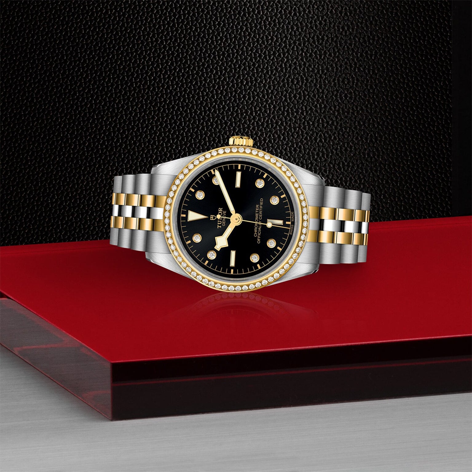 A TUDOR Black Bay One 36 S&G wristwatch in yellow gold and silver, featuring a black dial and gold bezel, is displayed on a red platform against a textured black background.