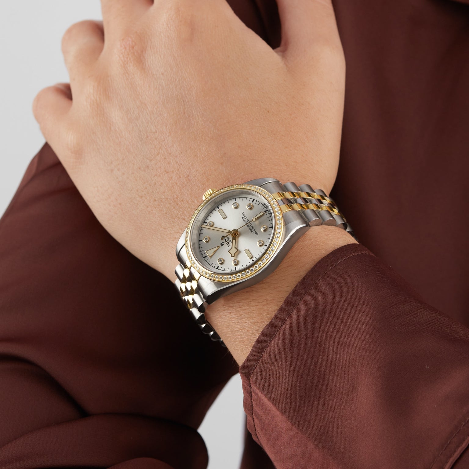 A person in a long-sleeve brown shirt wears a TUDOR Black Bay One 36 S&G two-tone gold and silver watch with a white dial and diamond hour markers.