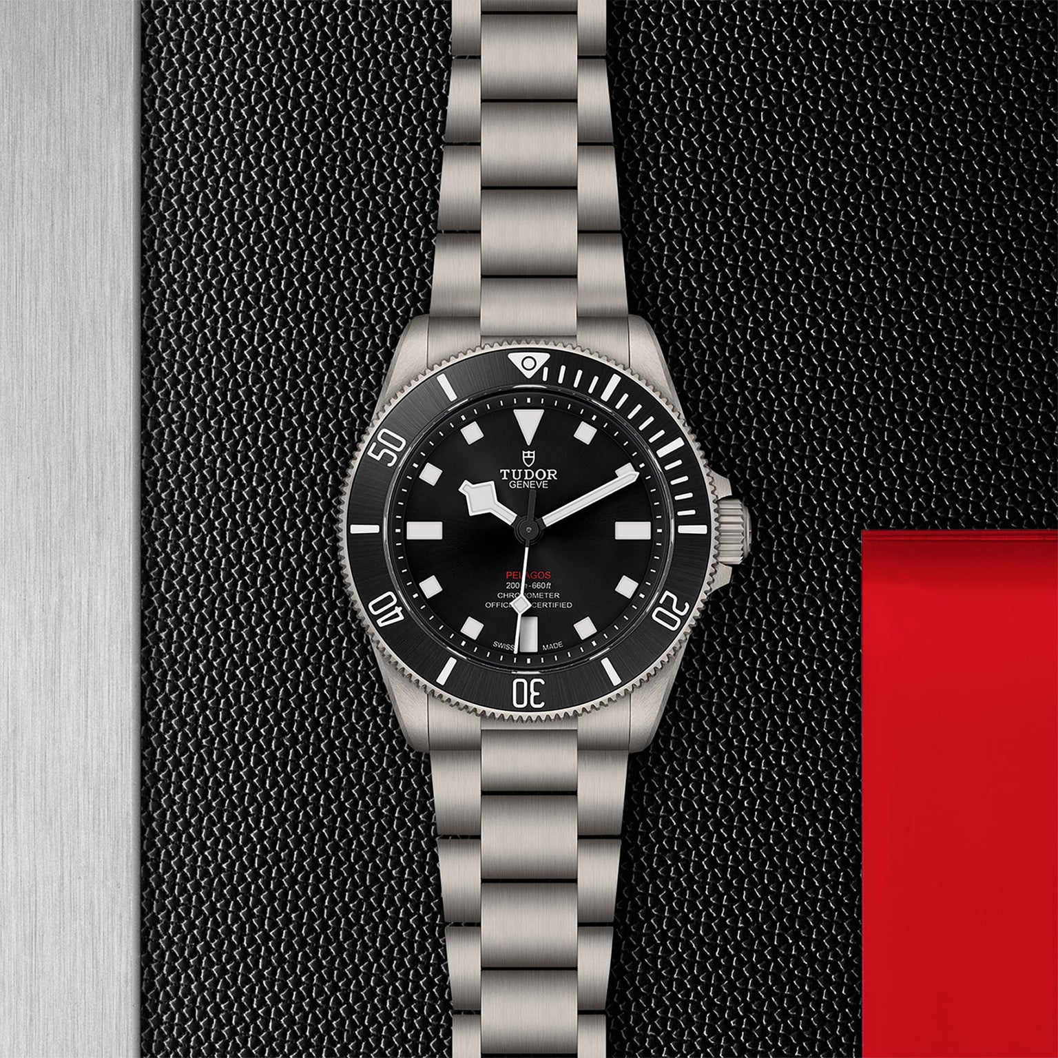 The TUDOR Pelagos 39, a stainless steel wristwatch with a black dial and bezel, is displayed on a textured black surface with metallic and red accents—an outstanding professional dive watch for mechanical watch enthusiasts.