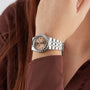 A person with long brown hair in a brown shirt displays the TUDOR Royal, a silver wristwatch with a pink dial, Roman numerals, and automatic movement—ideal for sport chic style.
