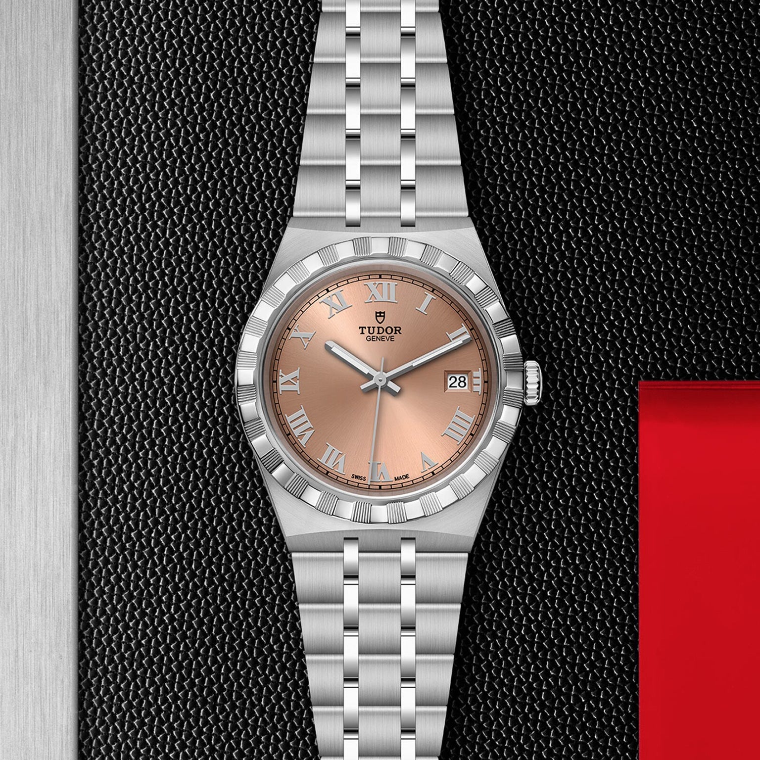 A TUDOR Royal wristwatch with a salmon dial, Roman numerals, date display, and stainless steel bracelet showcases sport chic style and automatic movement as it rests on a textured black surface.