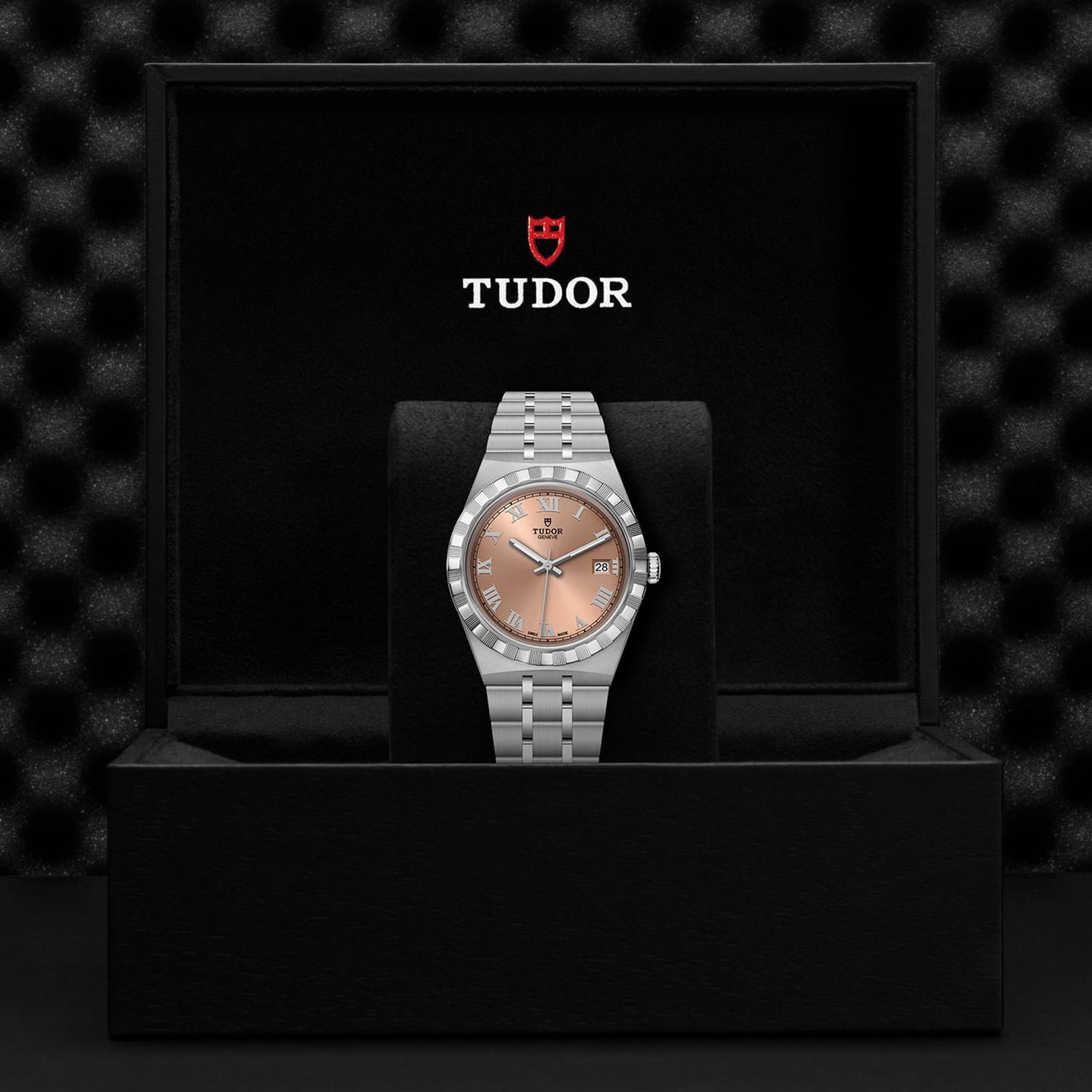 A TUDOR Royal wristwatch with a silver band and salmon dial, featuring automatic movement, is displayed upright in an open black presentation box branded with the TUDOR logo.