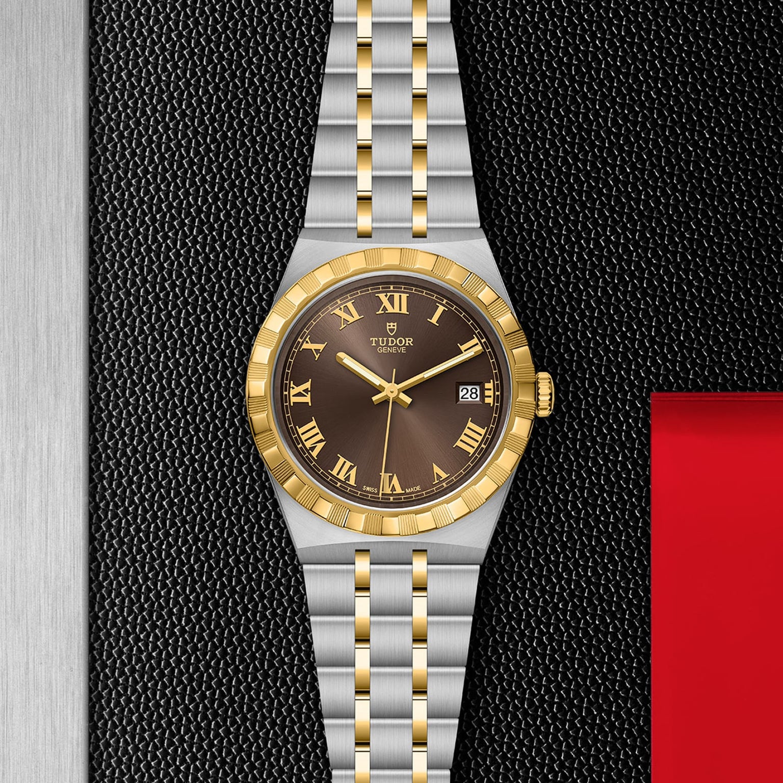 A sport-chic TUDOR Royal wristwatch with a gold and silver metal band, gold bezel, black dial with Roman numerals and date display, set against a textured black and metallic backdrop. Features reliable automatic movement.