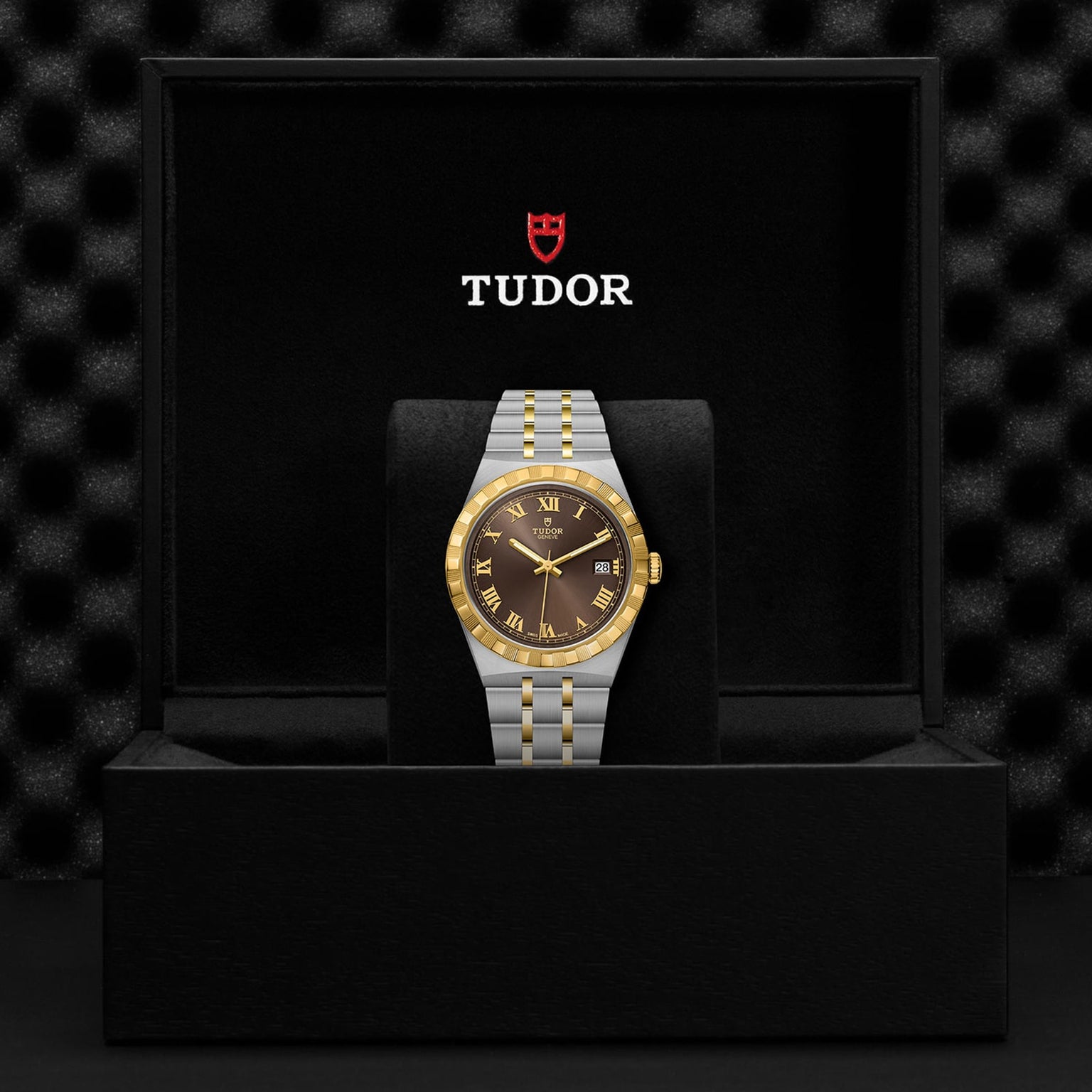 A TUDOR Royal wristwatch with a gold and silver band and a black and gold dial, featuring automatic movement, is showcased in an open black Tudor-branded box.