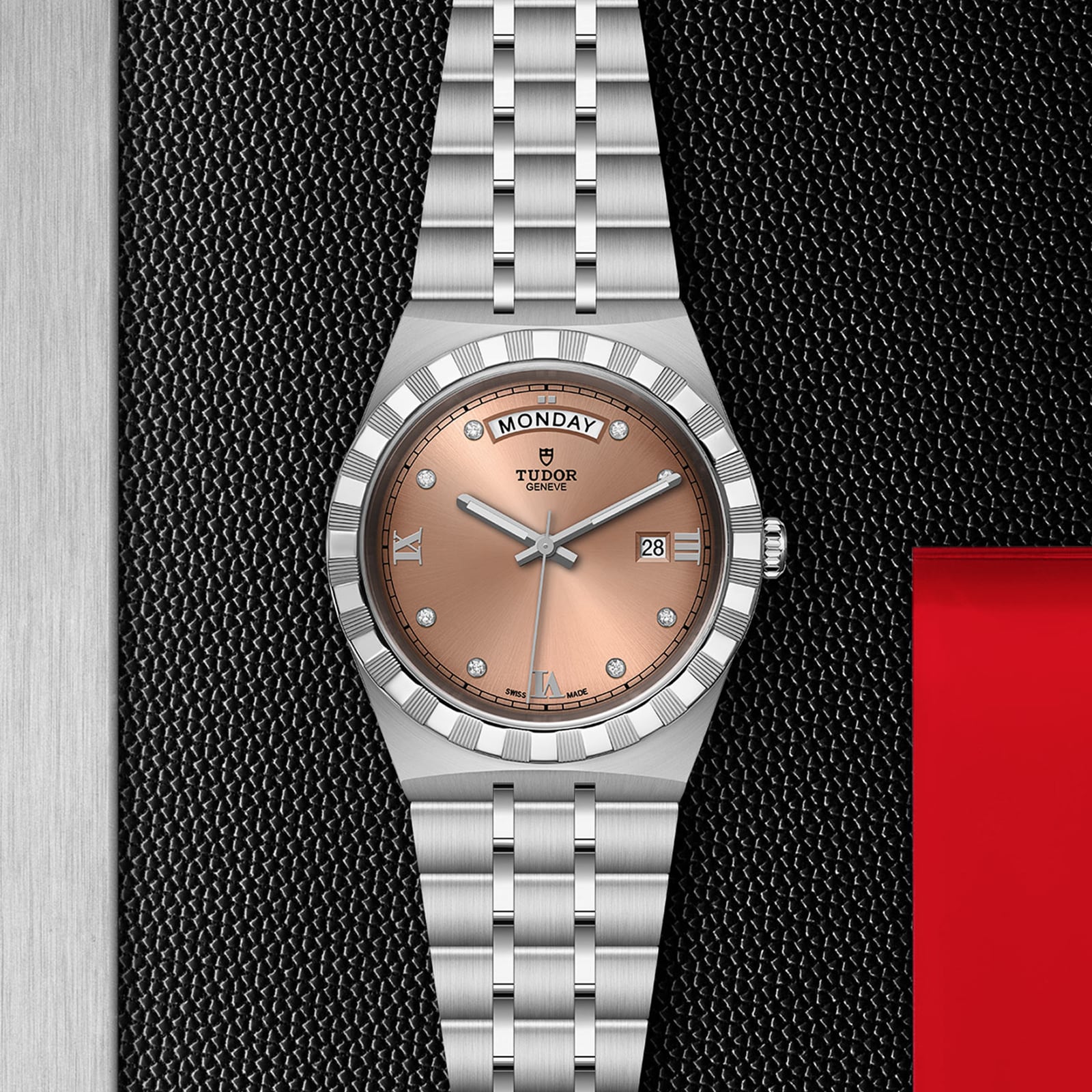 A TUDOR Royal wristwatch with a salmon dial, silver markers, day-date display, and stainless steel bracelet is shown on textured black, highlighting its sport-chic elegance.