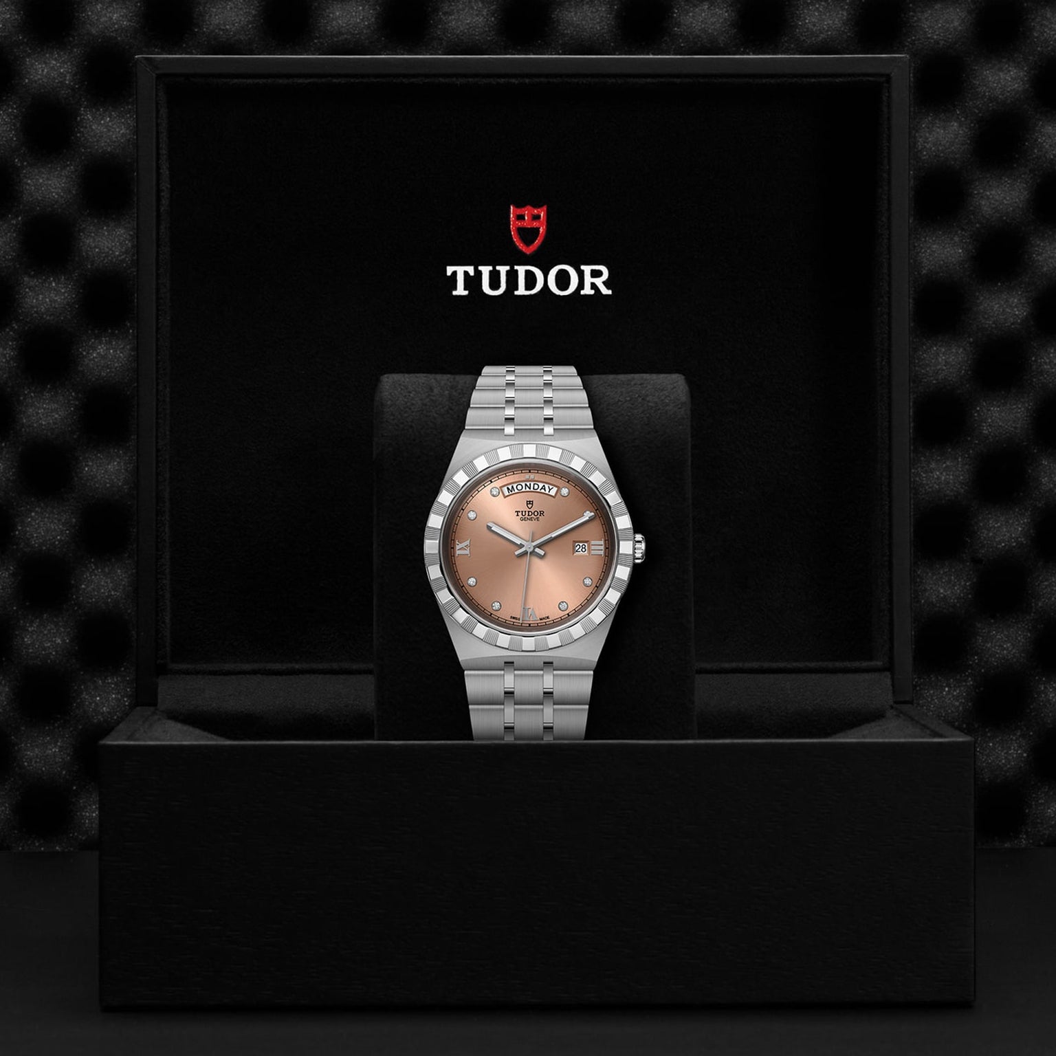 A TUDOR Royal wristwatch with a brown gradient dial, diamond-set bezel, and metal bracelet is displayed upright in a black box with the Tudor logo, highlighting the sophisticated sport-chic style of the TUDOR Royal.
