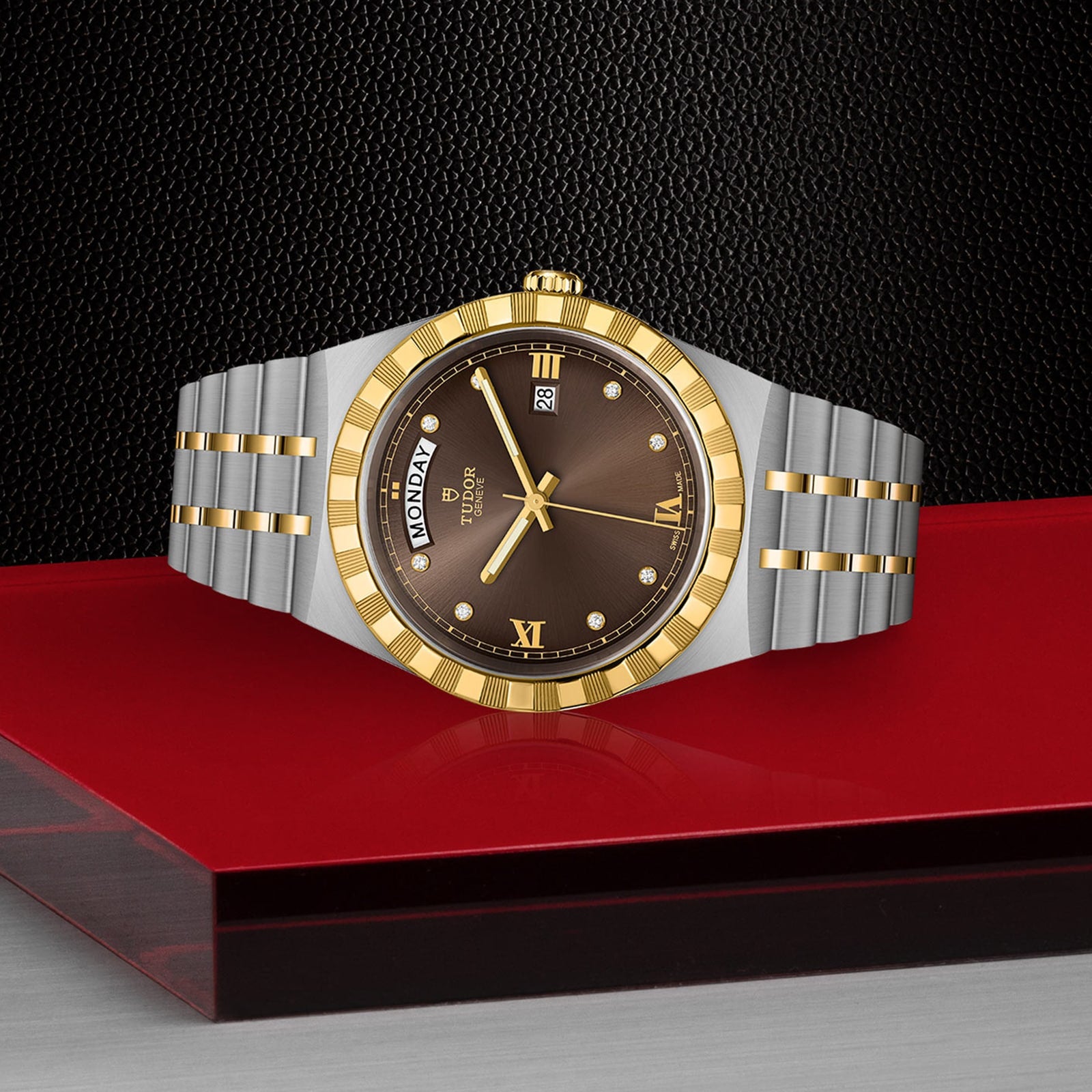 The TUDOR Royal wristwatch, featuring a gold bezel and brown dial, is showcased on a glossy red and black surface, exuding sport chic style against a textured black background.