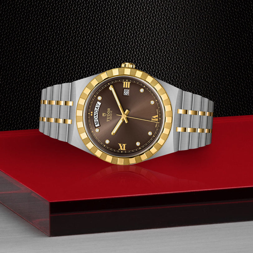 The TUDOR Royal wristwatch, featuring a gold bezel and brown dial, is showcased on a glossy red and black surface, exuding sport chic style against a textured black background.