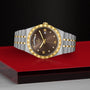 The TUDOR Royal wristwatch, featuring a gold bezel and brown dial, is showcased on a glossy red and black surface, exuding sport chic style against a textured black background.