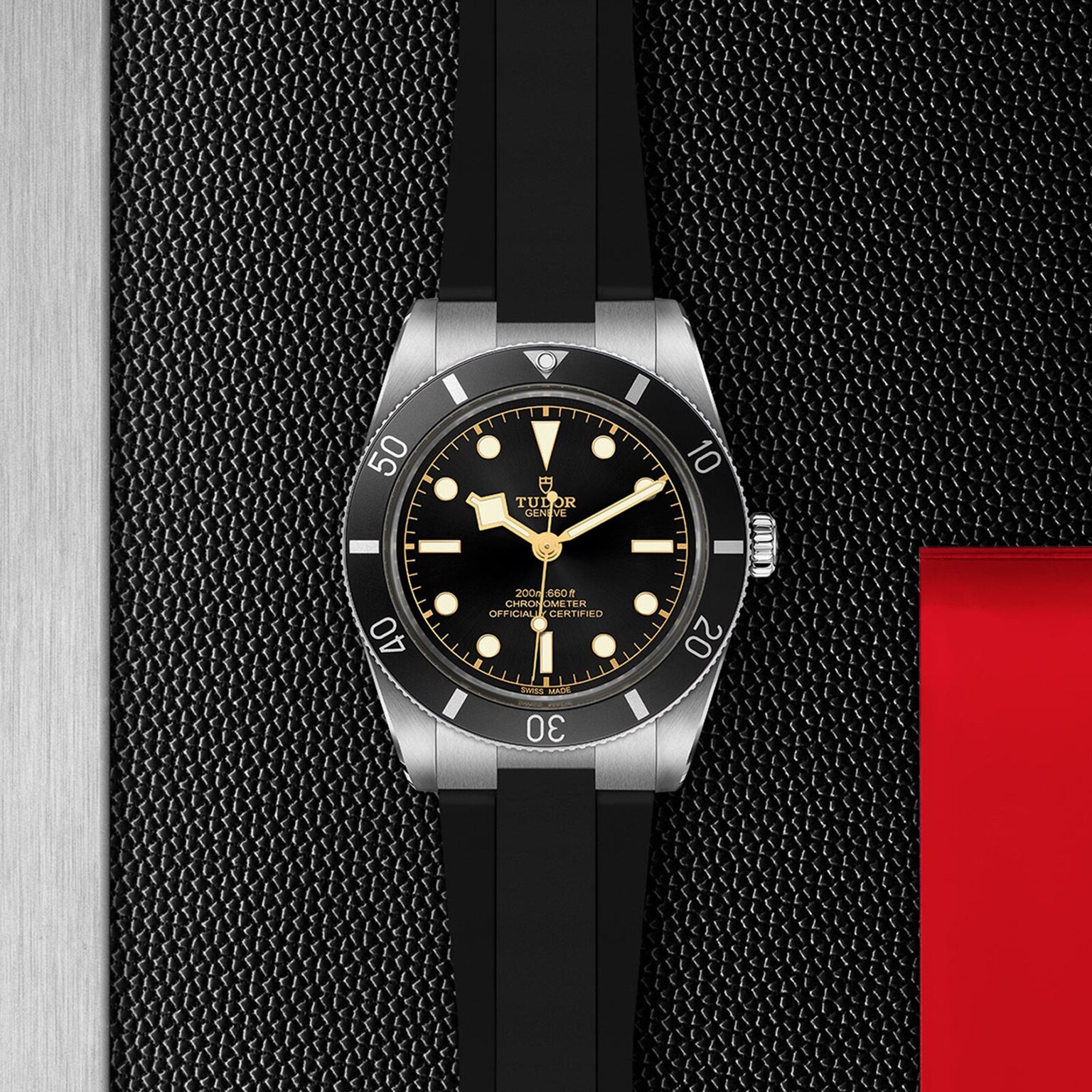 The TUDOR Black Bay 54 dive watch with a black dial, luminous markers, and a black strap is showcased on a textured black surface featuring metallic and red accents.