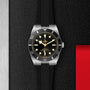The TUDOR Black Bay 54 dive watch with a black dial, luminous markers, and a black strap is showcased on a textured black surface featuring metallic and red accents.