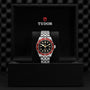 A TUDOR Black Bay wristwatch with a burgundy bezel and metal bracelet stands upright in a black presentation box with the Tudor logo above.