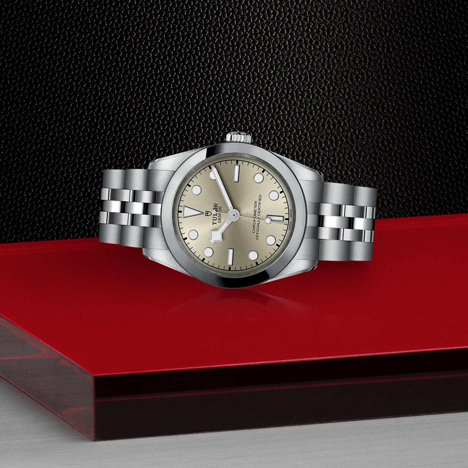 A TUDOR Black Bay One 31 in stainless steel with a champagne dial and bracelet is displayed on a glossy red and black surface, set against a textured black background.