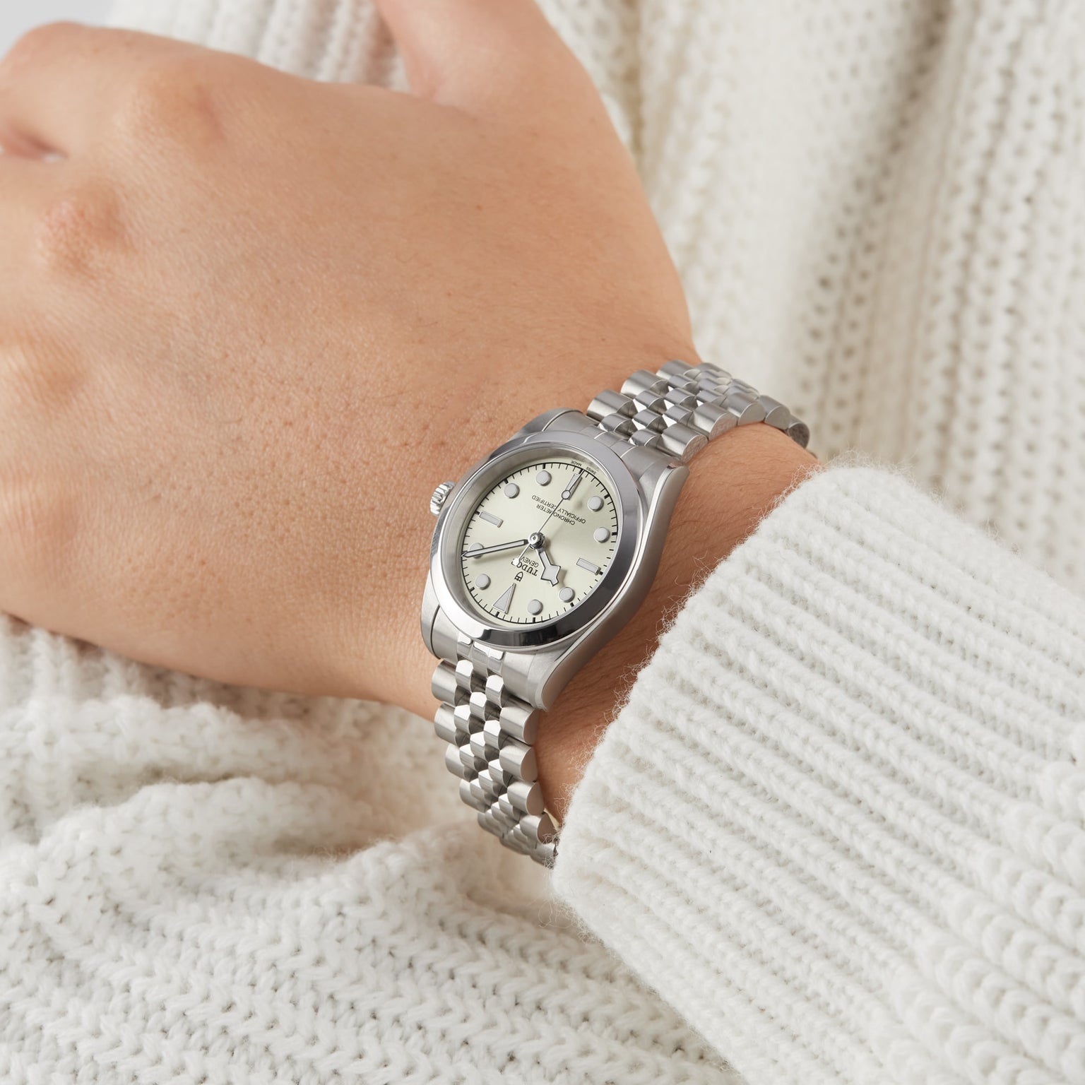 A TUDOR Black Bay One 31 wristwatch with a metal band on a persons wrist, paired with a white knitted sweater.