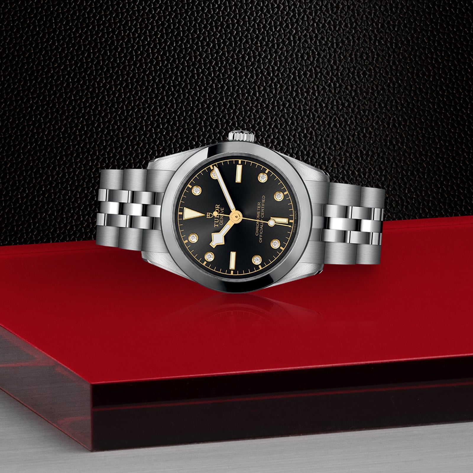 The TUDOR Black Bay 31 is a unisex chic stainless steel watch with a black dial and silver-gold hour markers, inspired by the refined Black Bay line and showcased on a bold red and black geometric background.