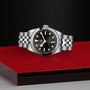 The TUDOR Black Bay 31 is a unisex chic stainless steel watch with a black dial and silver-gold hour markers, inspired by the refined Black Bay line and showcased on a bold red and black geometric background.