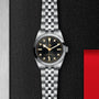 The TUDOR Black Bay 31 unisex wristwatch features a stainless steel case, black dial with gold hour markers, and a jubilee bracelet—shown here on an elegant black and metallic textured background.