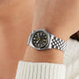 The TUDOR Black Bay 31 is a unisex silver wristwatch with a black dial and metal link band, shown worn over a cream-colored sweater.