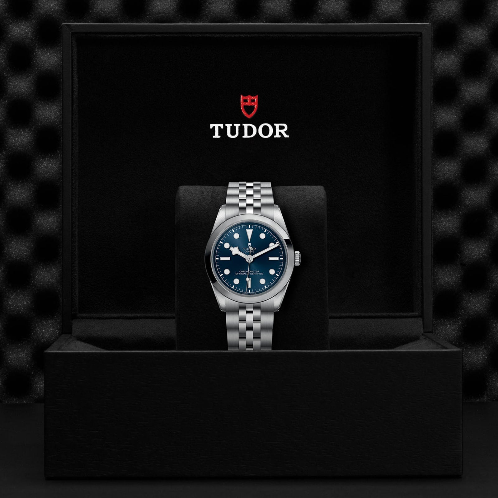A stylish TUDOR Black Bay One 36 stainless steel wristwatch with a blue dial is showcased on a cushion inside an open black box bearing the TUDOR logo.