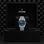 The TUDOR Black Bay One 36 wristwatch, featuring a blue dial, silver metal bracelet, and TUDOR rapid adjustment system, is displayed upright in an open black TUDOR-branded box.