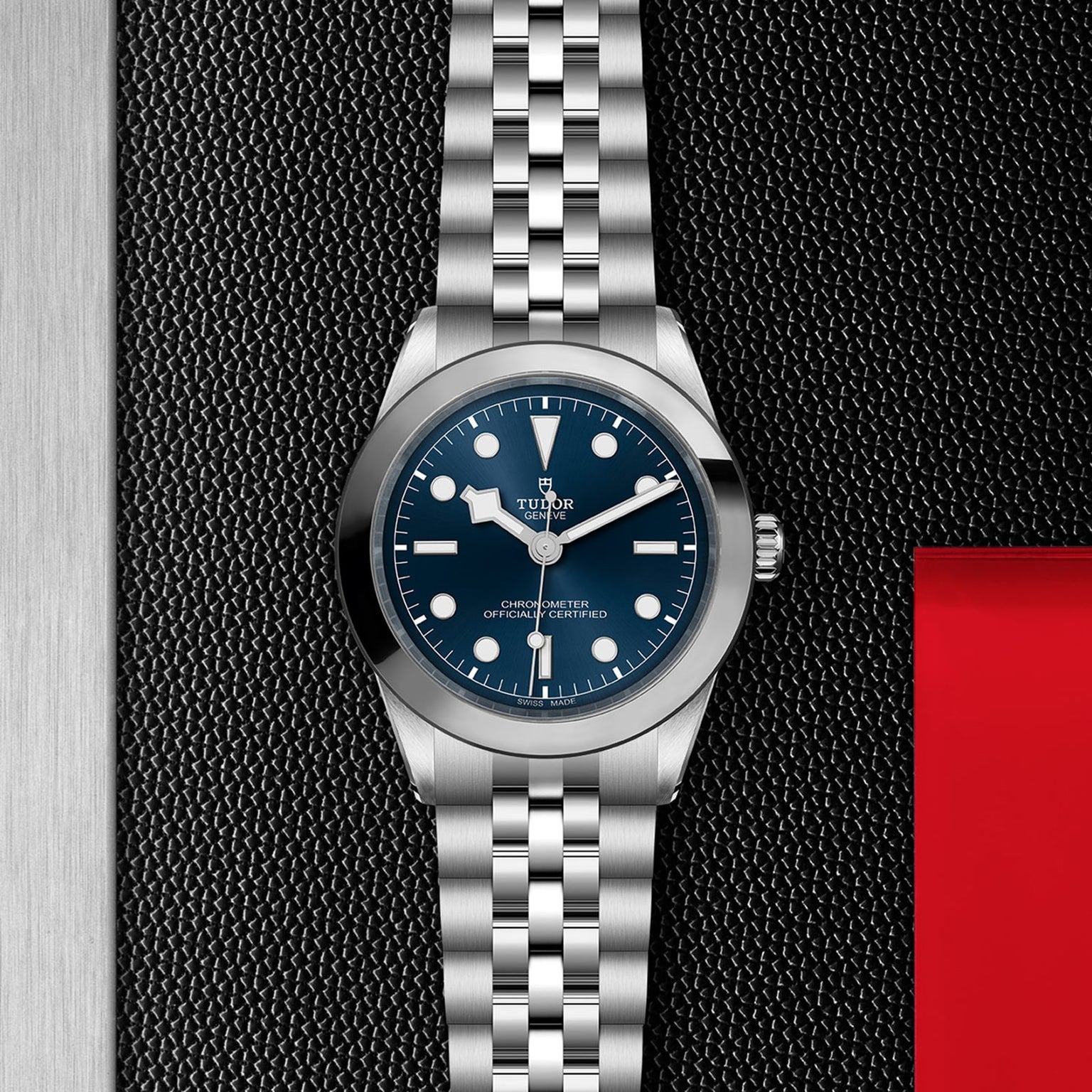 The TUDOR Black Bay One 39, a stainless steel wristwatch with a blue dial and silver bracelet inspired by Black Bay models, rests on a black textured surface accented with red and metallic highlights for unisex style.