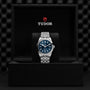 The TUDOR Black Bay One 39, a unisex wristwatch with a blue dial and silver metal band, is showcased upright in its open black box featuring the TUDOR logo, reflecting the refined Black Bay style and advanced Manufacture Calibres.
