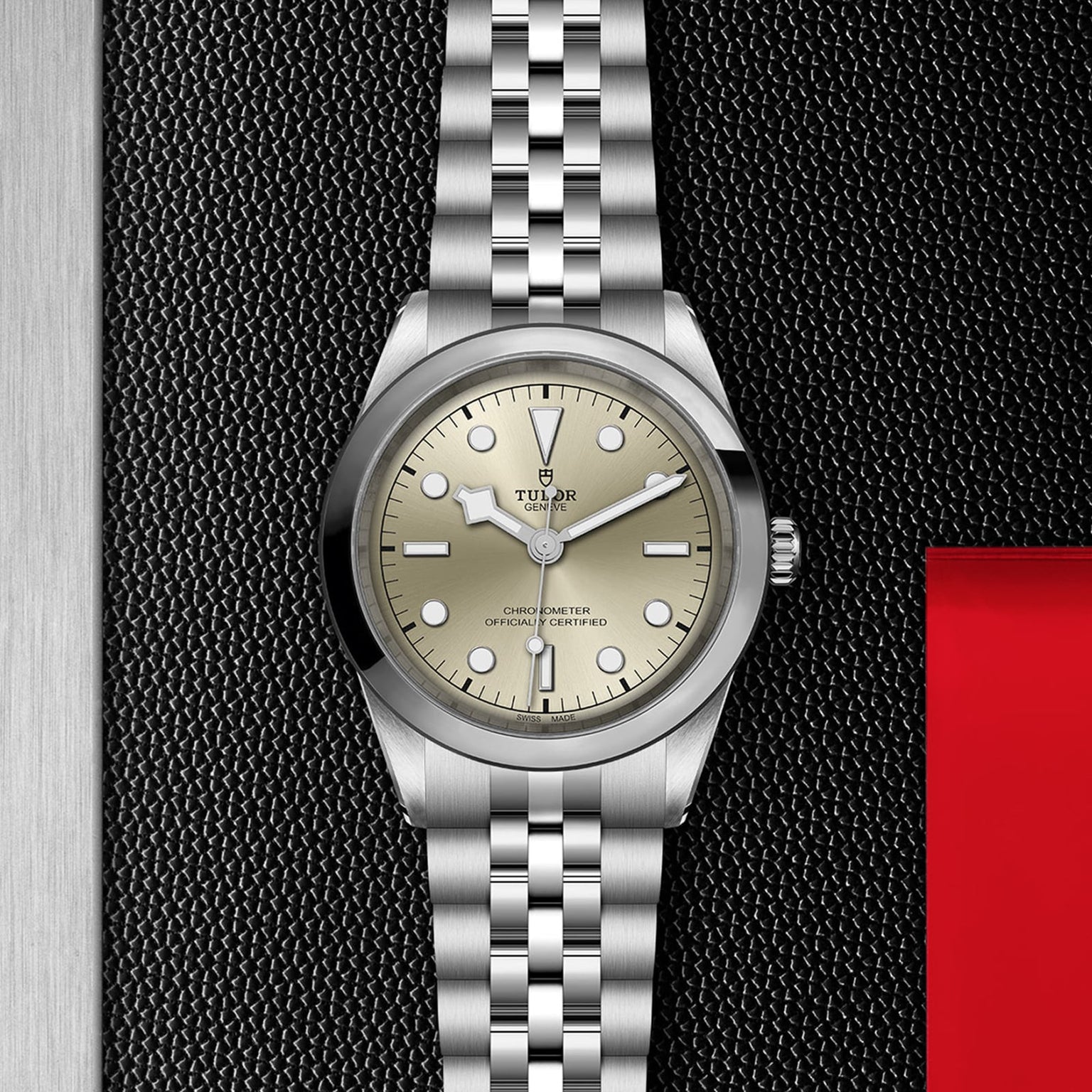 A TUDOR Black Bay One 41 wristwatch with a silver bracelet and champagne dial rests on a textured black surface, highlighting unisex chic with steel and red accents.