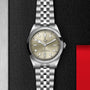 A TUDOR Black Bay One 41 wristwatch with a silver bracelet and champagne dial rests on a textured black surface, highlighting unisex chic with steel and red accents.