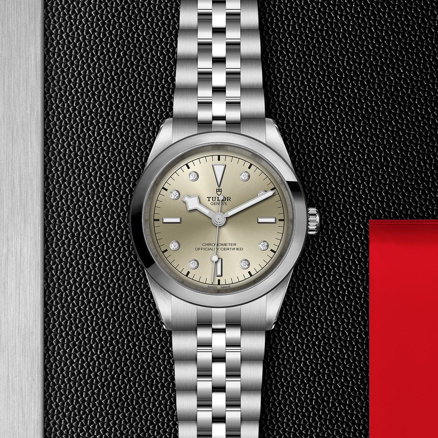 The TUDOR Black Bay One 41, a unisex silver wristwatch with a champagne dial, diamond hour markers, and metal bracelet, is displayed on a textured black surface beside an eye-catching red and silver border.