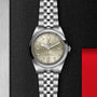 The TUDOR Black Bay One 41, a unisex silver wristwatch with a champagne dial, diamond hour markers, and metal bracelet, is displayed on a textured black surface beside an eye-catching red and silver border.